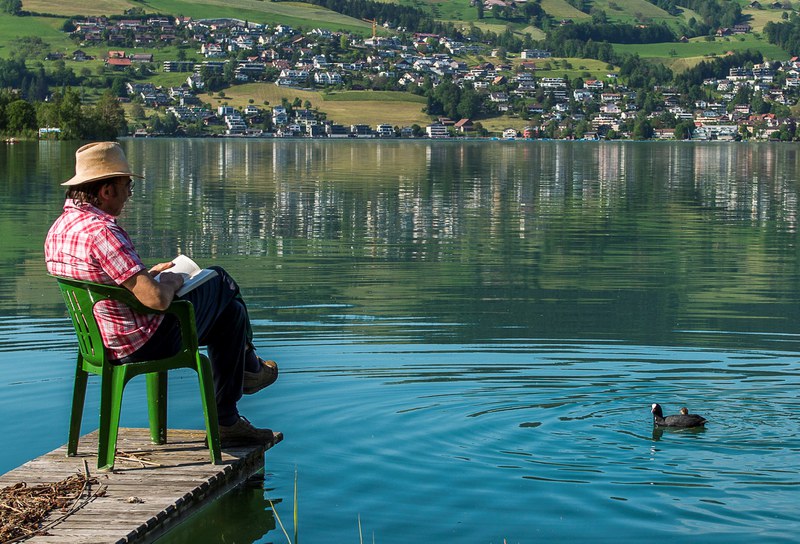 Mann liest auf einem See-Steg ein Buch in einem Stuhl